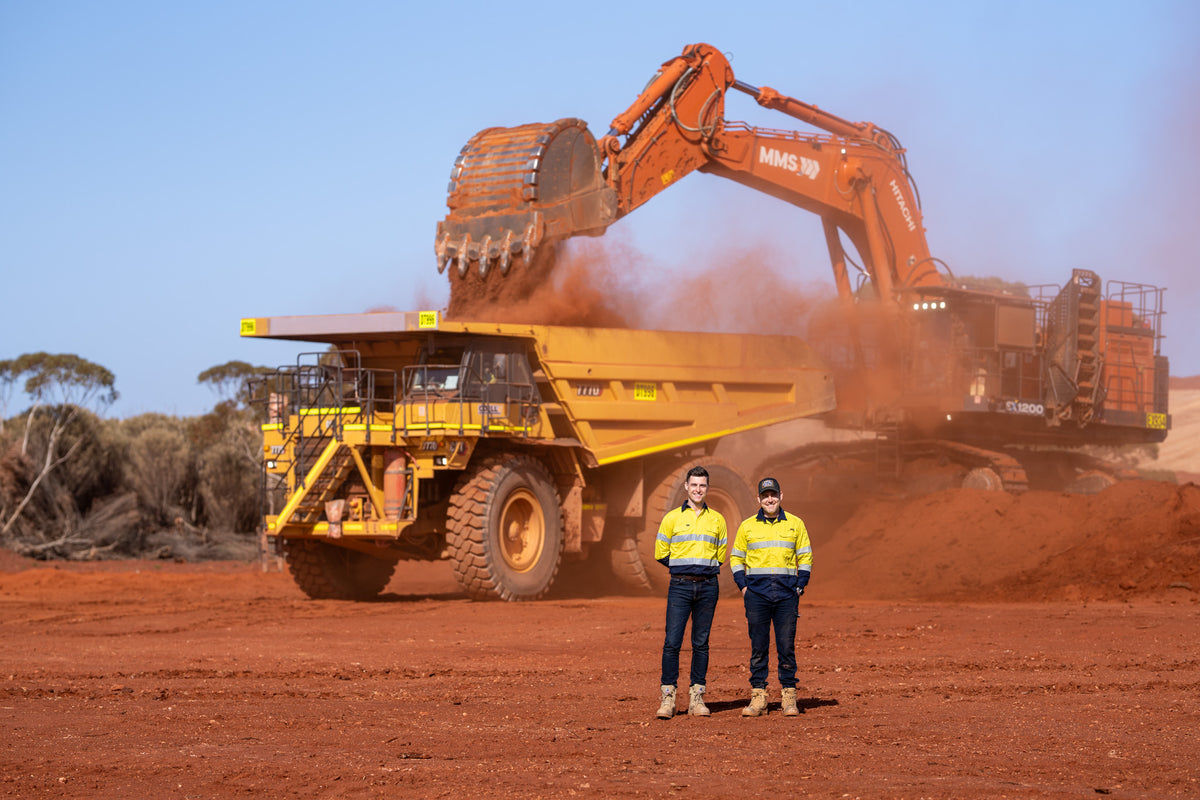 Two people in high-visibility workwear in front of a Hitachi excavator loading a mining haul truck